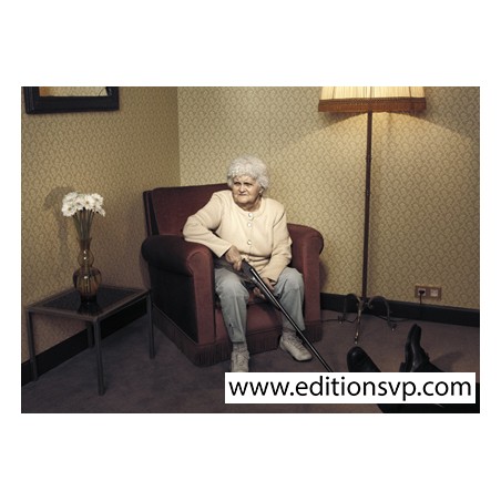 une femme agée assise dans un fauteuil tient un fusil de chasse, au sol on voit les pieds d'un homme allongé,